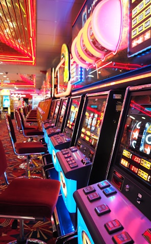 A brightly lit casino interior featuring a row of colorful slot machines. The neon lights create a vibrant atmosphere, with patterned carpeting adding to the decor. Stools are aligned neatly in front of each machine.