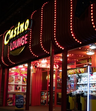 A brightly lit casino lounge entrance adorned with neon lights. The exterior features colorful signage including the words 'Casino' and 'Lounge' in vivid colors, with additional lights framing the building. Inside, a glimpse of a claw machine filled with plush toys and arcade games is visible. The area exudes an inviting yet bustling atmosphere typical of entertainment venues.