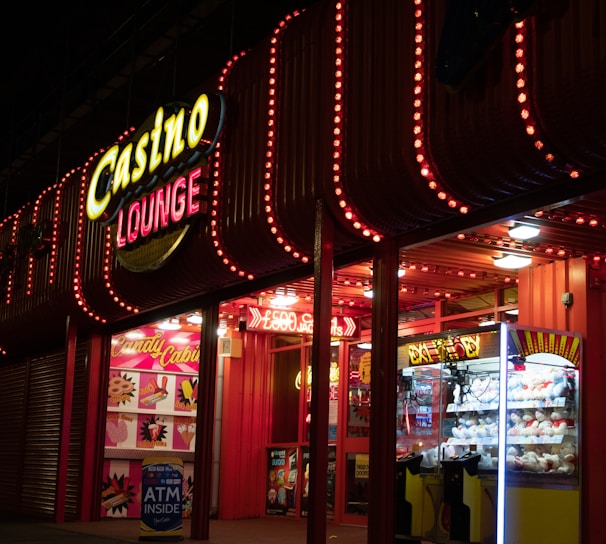 A brightly lit casino lounge entrance adorned with neon lights. The exterior features colorful signage including the words 'Casino' and 'Lounge' in vivid colors, with additional lights framing the building. Inside, a glimpse of a claw machine filled with plush toys and arcade games is visible. The area exudes an inviting yet bustling atmosphere typical of entertainment venues.